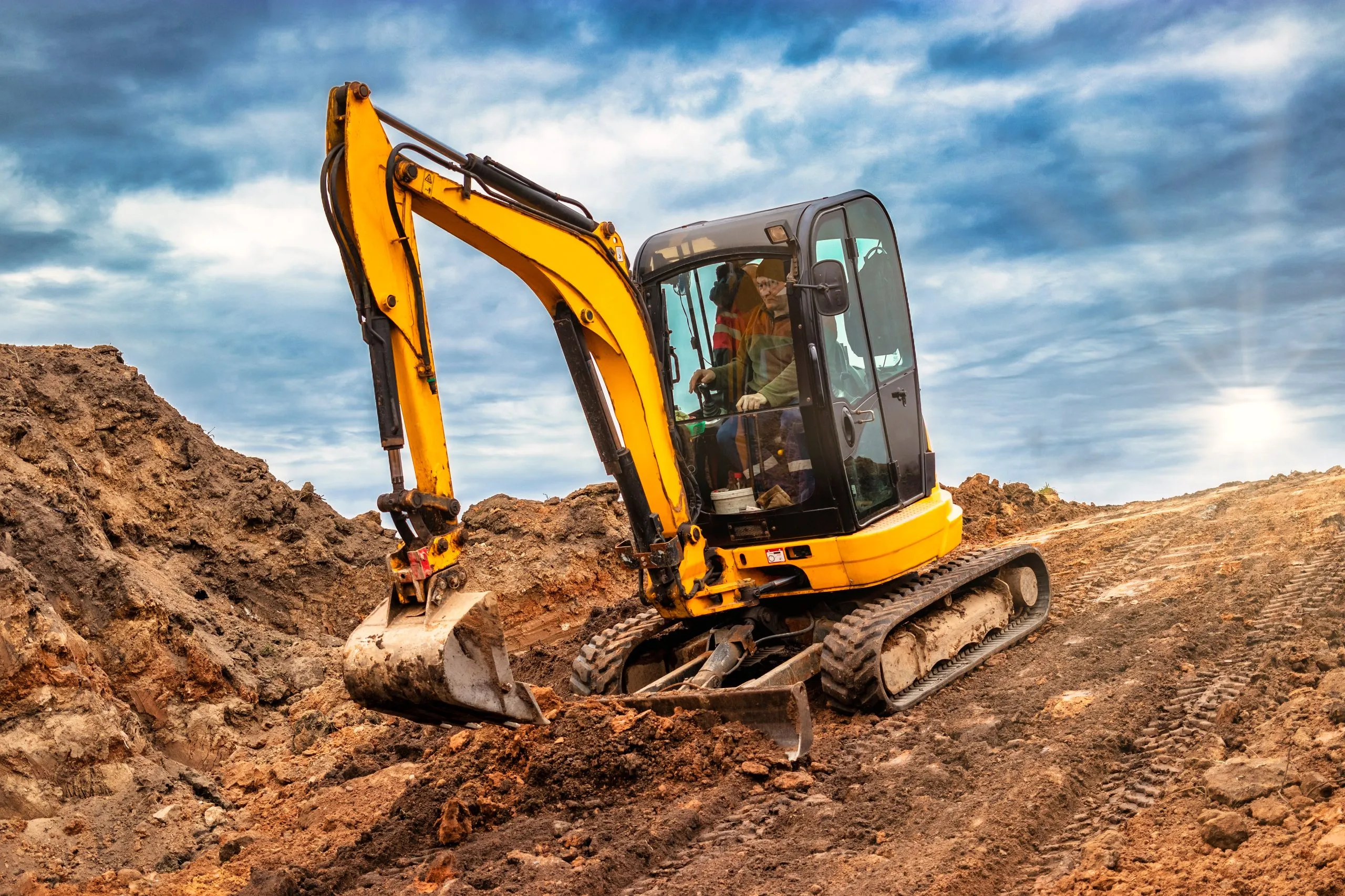 Yellow mini excavator with operator working on a dirt mound under a sunny sky