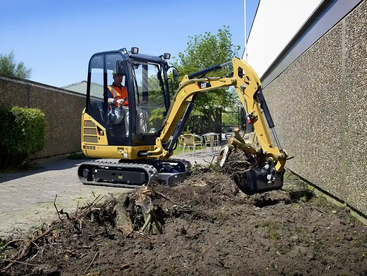 Mini excavator with operator clearing roots and soil beside a building wall